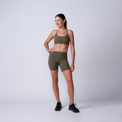 Smiling woman wears olive green Women’s Biker Shorts - ToughFlex and black sneakers, standing against a plain light gray background.
