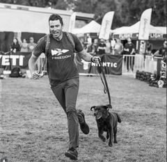 A man wearing the Freedom Tee - Tri-Blend runs with a smiling dog on a leash at an outdoor GORUCK event; both are happy, captured in black and white.