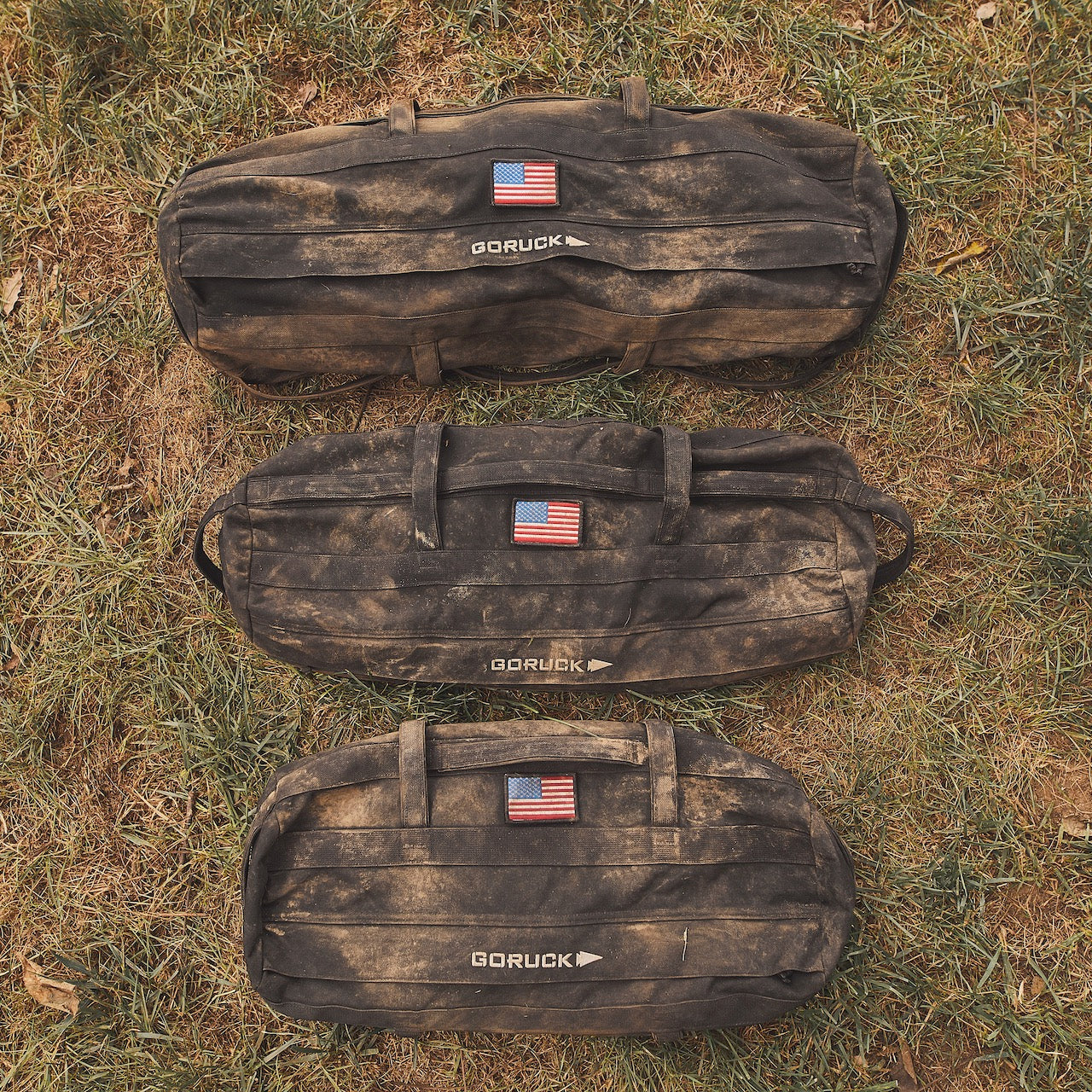 Three rugged Sandbag Kits (w/ Filler Bag), each featuring an American flag patch, rest on the grass—built tough and ready to power your most demanding workouts.