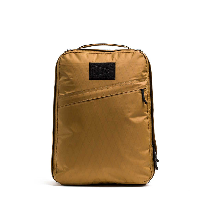 The GR0 - X-PAC is a tan, rectangular, lightweight rucksack made from waterproof material, featuring a front zipper pocket and a black patch logo.