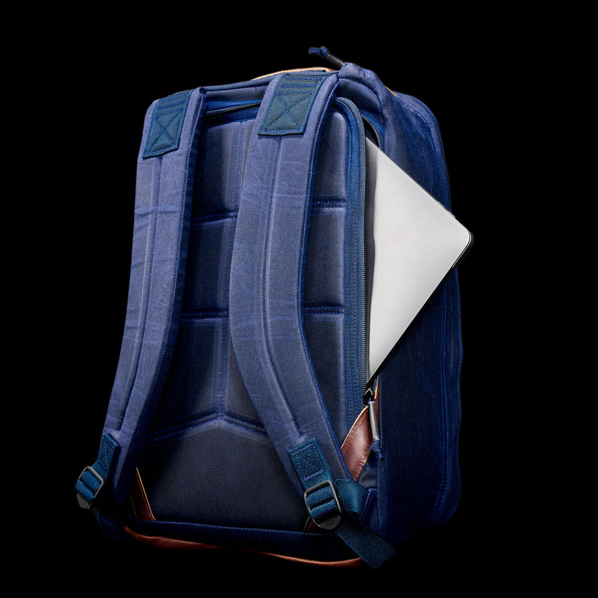 The GR1 USA - Heritage Waxed Canvas backpack features padded straps and a partially visible laptop in its back pocket, displayed against a black background.