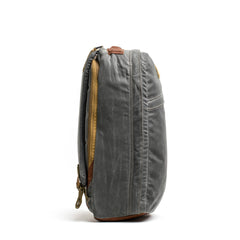 A side view of the GR1 USA - Heritage rucksack by GORUCK, featuring gray waxed canvas with brown leather accents and a zipper. The fabric showcases a rugged texture against the plain white background.