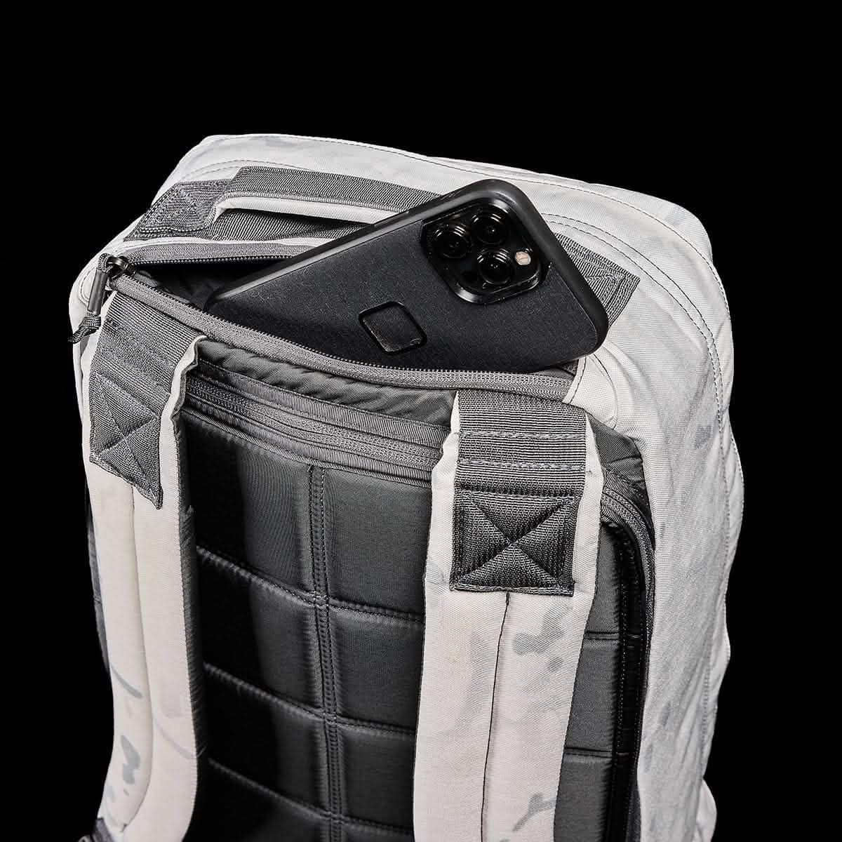 The GR1 backpack, with a phone partially tucked into its zippered pocket, is shown against a black background, highlighting its rugged Special Forces–inspired design.
