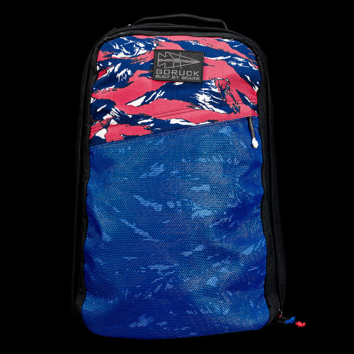 A blue and red GR1 Shooter 26L - Built by Scars backpack, crafted from rugged CORDURA® fabric with a mountain pattern and black zipper, is displayed against a black background.