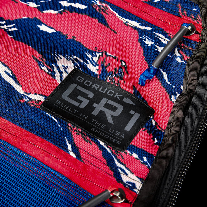 Close-up of a GR1 Shooter 26L - Built by SCARS label on a vibrant CORDURA® fabric bag in red, blue, and white with visible zippers—showcasing durability from the SCARS team.