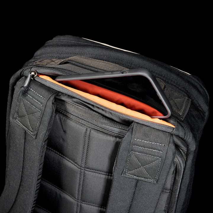 A tablet partially slides out of an orange-lined pocket in the black GR1 Shooter 26L - Built by scars backpack, made from durable CORDURA®, set against a black background.