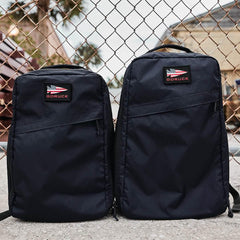 Two black GORUCK rucksacks with American flag patches side by side, against a chain-link fence background