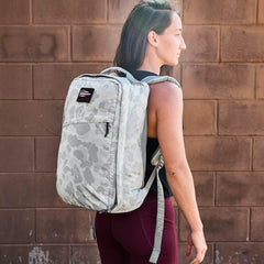 Woman wearing light gray camouflage GORUCK backpack standing against brown brick wall