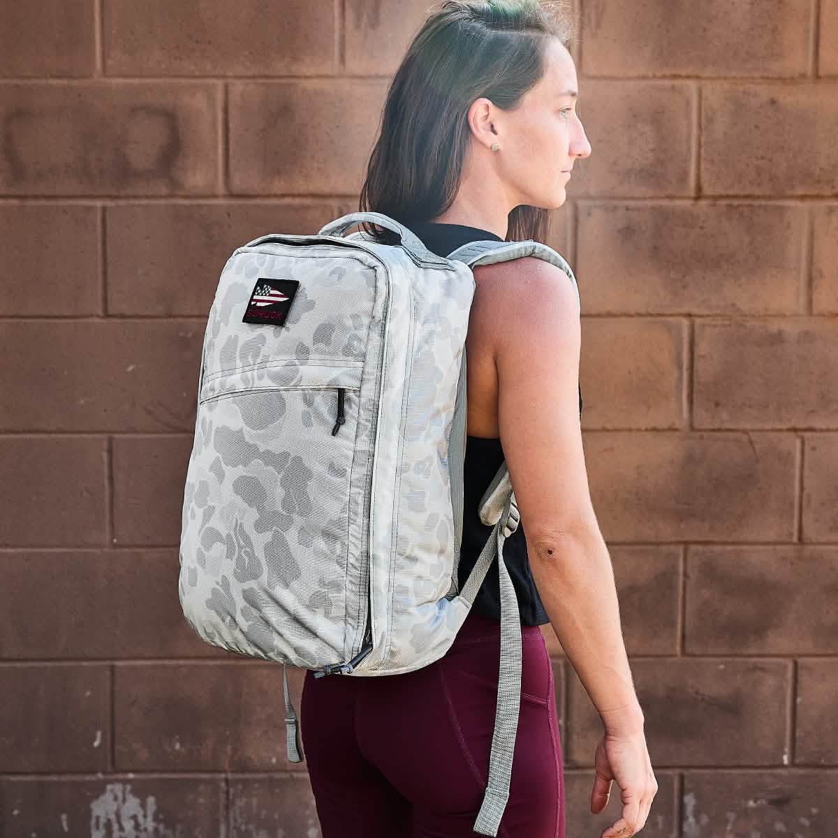 A person with long hair stands against a brown brick wall, wearing burgundy leggings and the ultra-lightweight GR1 USA - Ripstop ROBIC® rucksack by GORUCK, which features a front pocket and adjustable straps.
