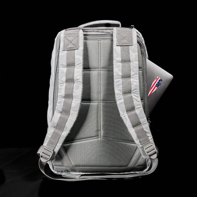 A gray GR1 USA - Ripstop ROBIC® backpack from GORUCK stands upright, showcasing its water-resistant design and padded straps, with a laptop partially visible in the side pocket adorned with an American flag sticker. The black background highlights the rucksack's sleek design.