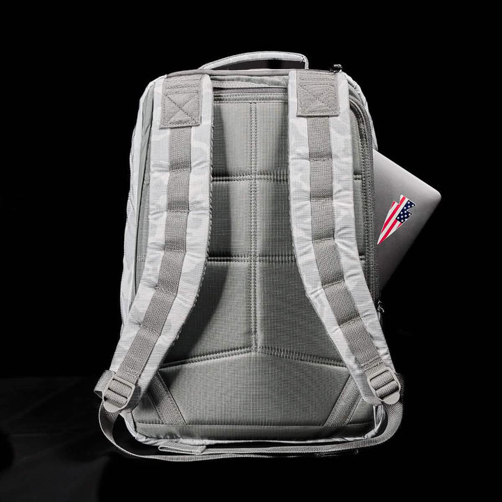 A gray GR1 USA - Ripstop ROBIC® backpack from GORUCK stands upright, showcasing its water-resistant design and padded straps, with a laptop partially visible in the side pocket adorned with an American flag sticker. The black background highlights the rucksack's sleek design.