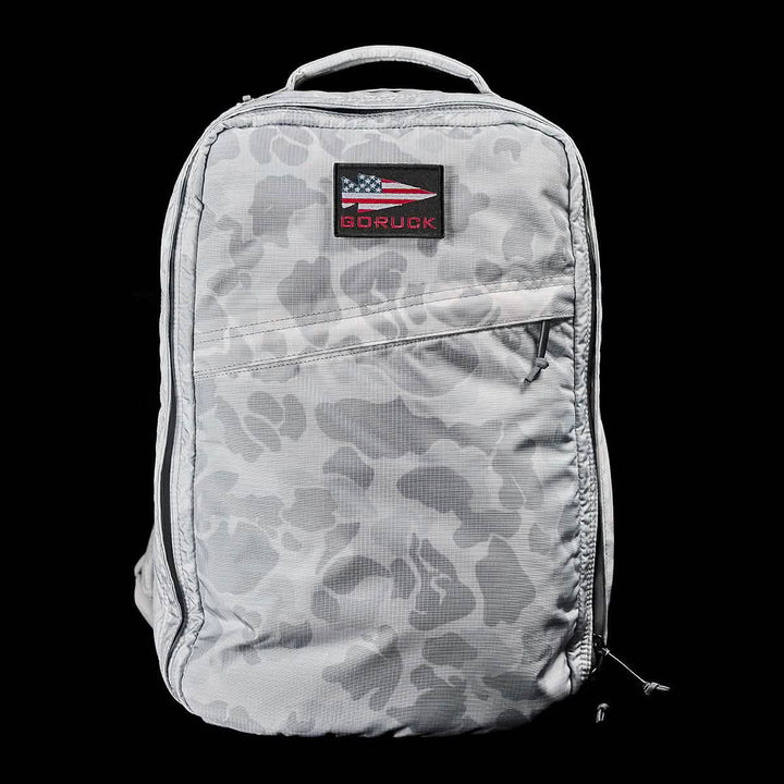 Introducing the GR1 USA - Ripstop ROBIC® by GORUCK, a gray camo-patterned rucksack equipped with a handy handle and a convenient front zippered pocket. It includes a distinctive black patch adorned with an American flag design and the word "GORUCK" in striking red. This ultra-lightweight and water-resistant backpack is engineered for any adventure, set against a sleek black background.