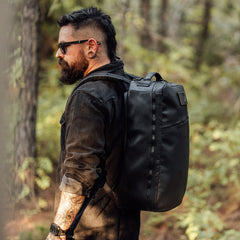 Tattooed bearded man with sunglasses stands in a forest, wearing the GR1 USA x Carryology - Berserker Viking backpack.