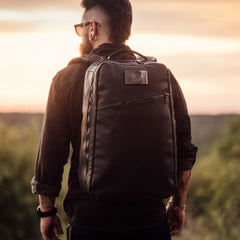 Bearded man in sunglasses wearing a black GR1 USA x Carryology - Berserker Viking backpack, standing outdoors at sunset.