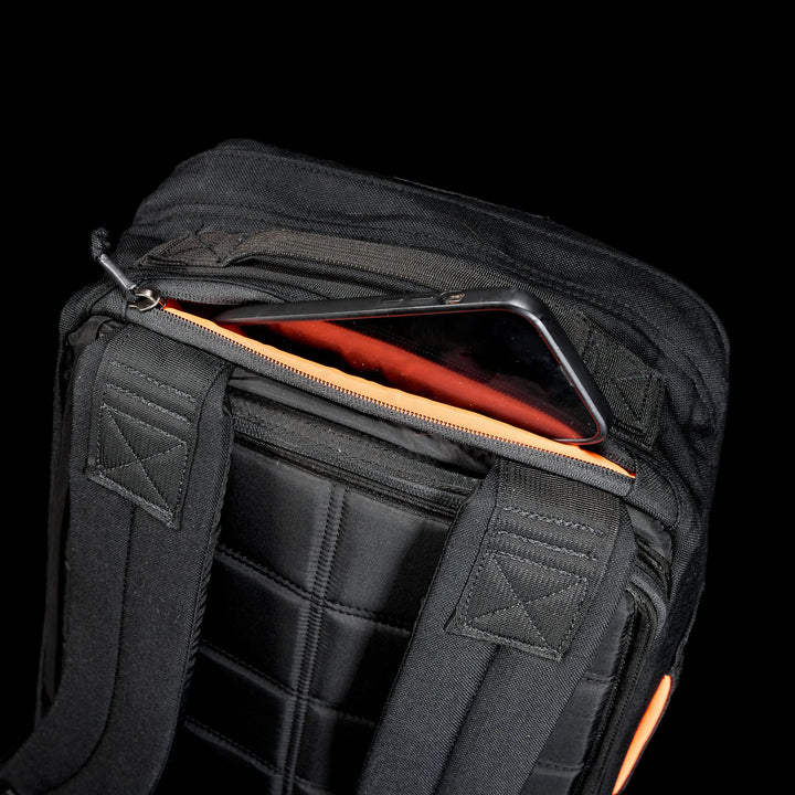 A smartphone partially tucked in a zippered pocket on a black GR1 Shooter 26L - Built by Scars backpack, crafted from Special Forces Grade CORDURA® with padded straps.