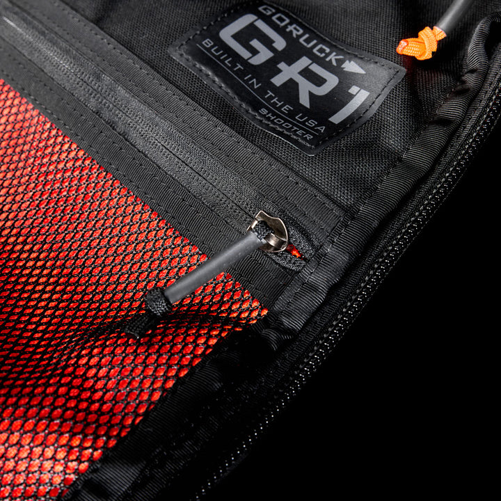 Close-up of a GR1 Shooter 26L - Built by Scars backpack pocket, made from rugged CORDURA®, featuring red mesh and a visible zipper pull.