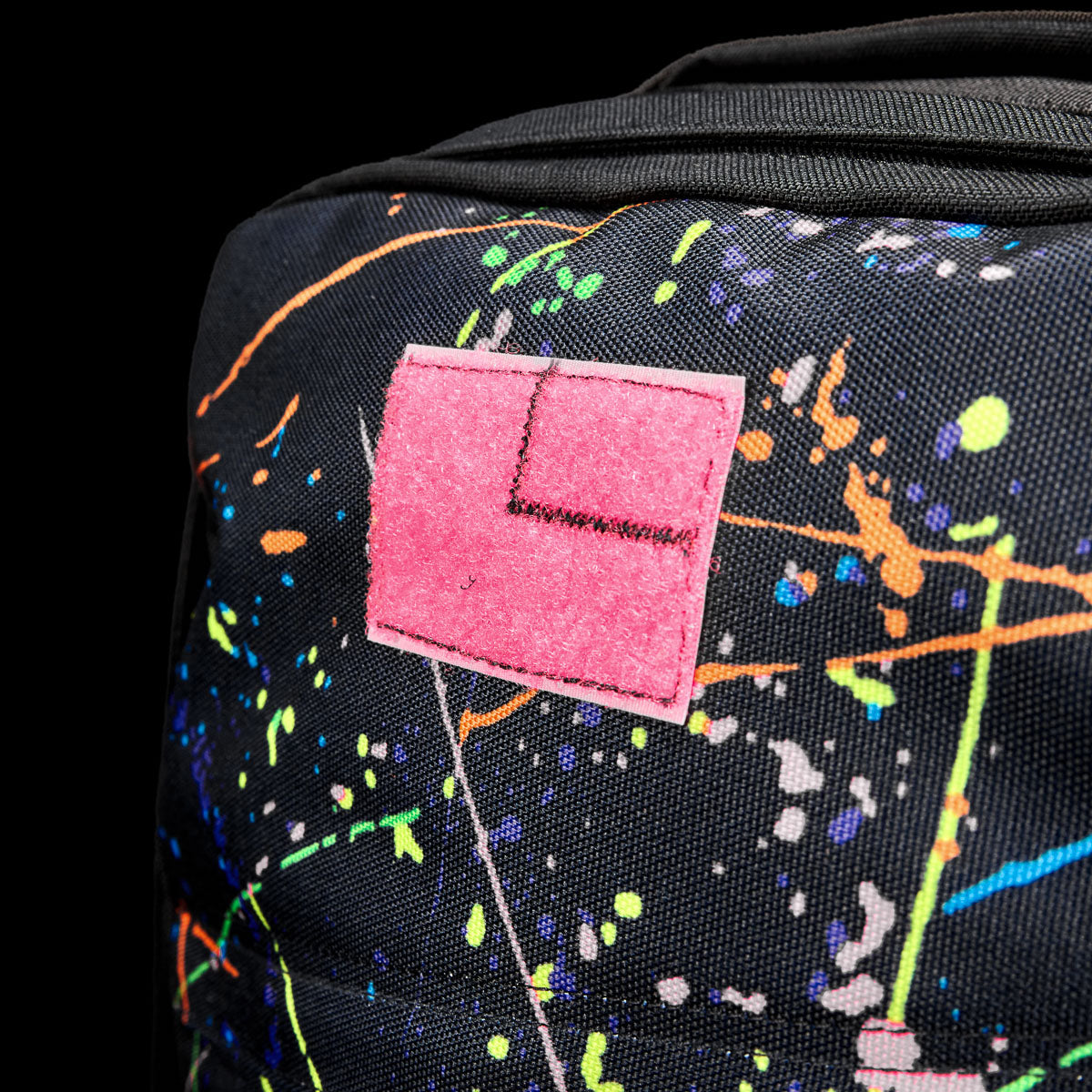 Close-up of the GR1 Shooter 26L - Built by SCARS in black, crafted from Special Forces Grade CORDURA®, accented with colorful paint splatters and a pink patch with an embroidered "L," reflecting its durability and style.