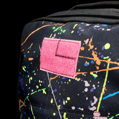 Close-up of the GR1 Shooter 26L - Built by SCARS in black, crafted from Special Forces Grade CORDURA®, accented with colorful paint splatters and a pink patch with an embroidered "L," reflecting its durability and style.