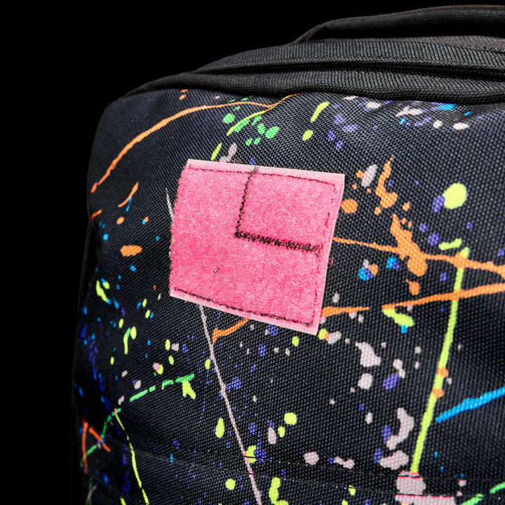 Close-up of the GR1 Shooter 26L - Built by SCARS in black, crafted from Special Forces Grade CORDURA®, accented with colorful paint splatters and a pink patch with an embroidered "L," reflecting its durability and style.