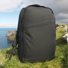 A black GR1 USA Slick - Ballistic Nylon Cordura backpack with a bombproof laptop compartment sits on grassy cliffs overlooking the ocean under a partly cloudy sky.