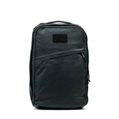 The GR1 USA - Heritage Waxed Canvas is a black, rectangular backpack featuring a top handle and a front diagonal zipper pocket.