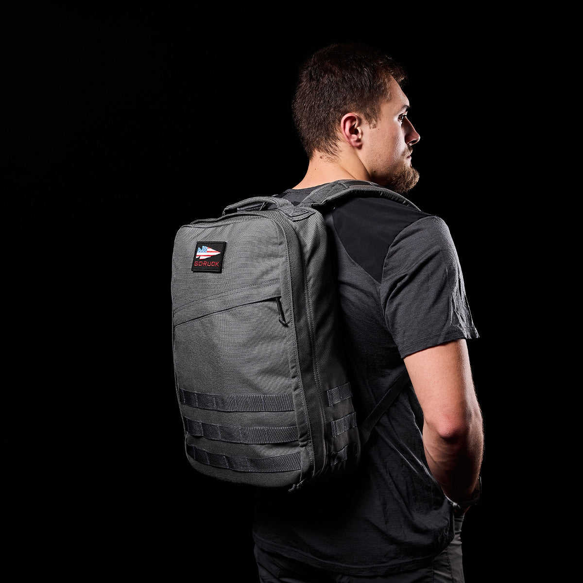 Man in a black shirt wearing a gray GR1 USA - Cordura rucksack, Special Forces tested for durability, standing against a black background.