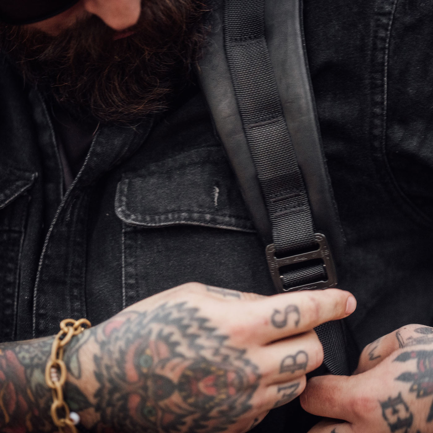 Tattooed person wearing a black denim shirt adjusts the strap of a black backpack.