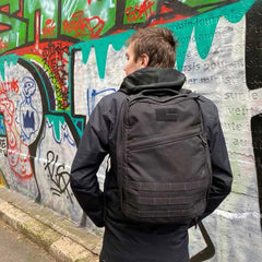 A person with a GR1 USA black backpack by GORUCK, celebrated for its American craftsmanship, stands facing a graffiti-covered wall.