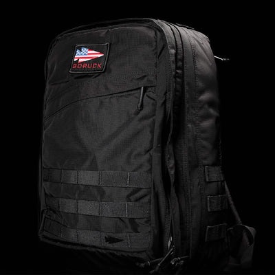 A GR2 - Ripstop ROBIC® by GORUCK in black, featuring multiple compartments and a patch with an American flag and the text GORUCK on the front. The dark background accentuates its sleek, water-resistant design and rugged texture, making it ideal for any adventure.