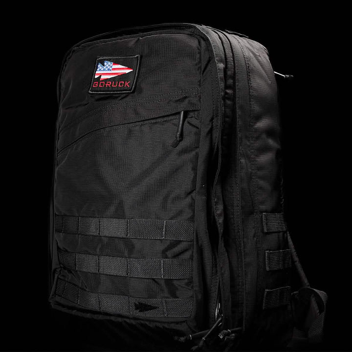 A GR2 - Ripstop ROBIC® by GORUCK in black, featuring multiple compartments and a patch with an American flag and the text GORUCK on the front. The dark background accentuates its sleek, water-resistant design and rugged texture, making it ideal for any adventure.
