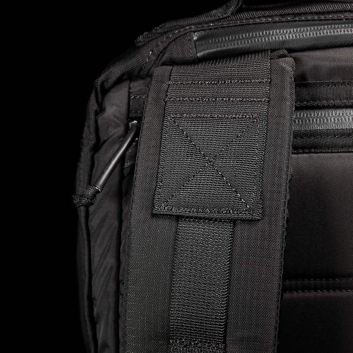 Close-up of the GR2 - Ripstop ROBIC® rucksack by GORUCK displaying detailed stitching and a padded strap. The texture of the fabric stands out, highlighting its water-resistant quality, with a partially visible metal clip. The dark background underscores the premium design and craftsmanship of this black travel bag.