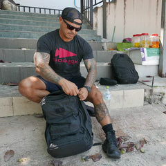 A person with tattoos sits on outdoor steps, wearing a black cap, sunglasses, and a gray T-shirt. They pack a GORUCK GR2 backpack, renowned for its Special Forces quality. Nearby are another carry-on compliant bag and a bottle of water amidst scattered fallen leaves.
