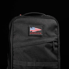 Set against a dark backdrop, the stylish GR2 backpack by GORUCK features a patch with an American flag and 'GORUCK' in red. Designed with Special Forces quality and a robust top handle, it ensures perfect carry-on compliance.