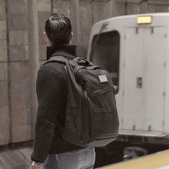 A person with a GORUCK GR2 backpack, celebrated for its Special Forces quality and adorned with an American flag patch, stands on a subway platform. A white train is approaching the underground station, recognized by its tiled walls.