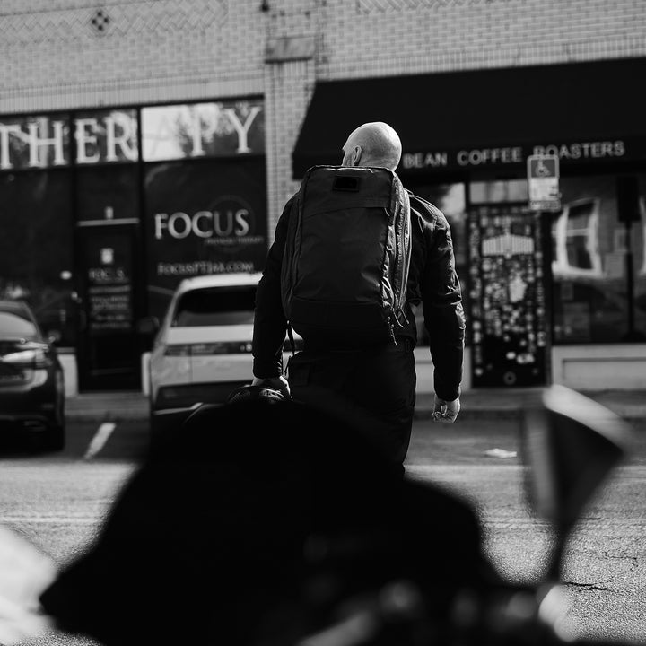 A bald man wearing the GR2 X-PAC backpack with AquaGuard zippers walks toward a coffee shop on a city street, seen from behind.