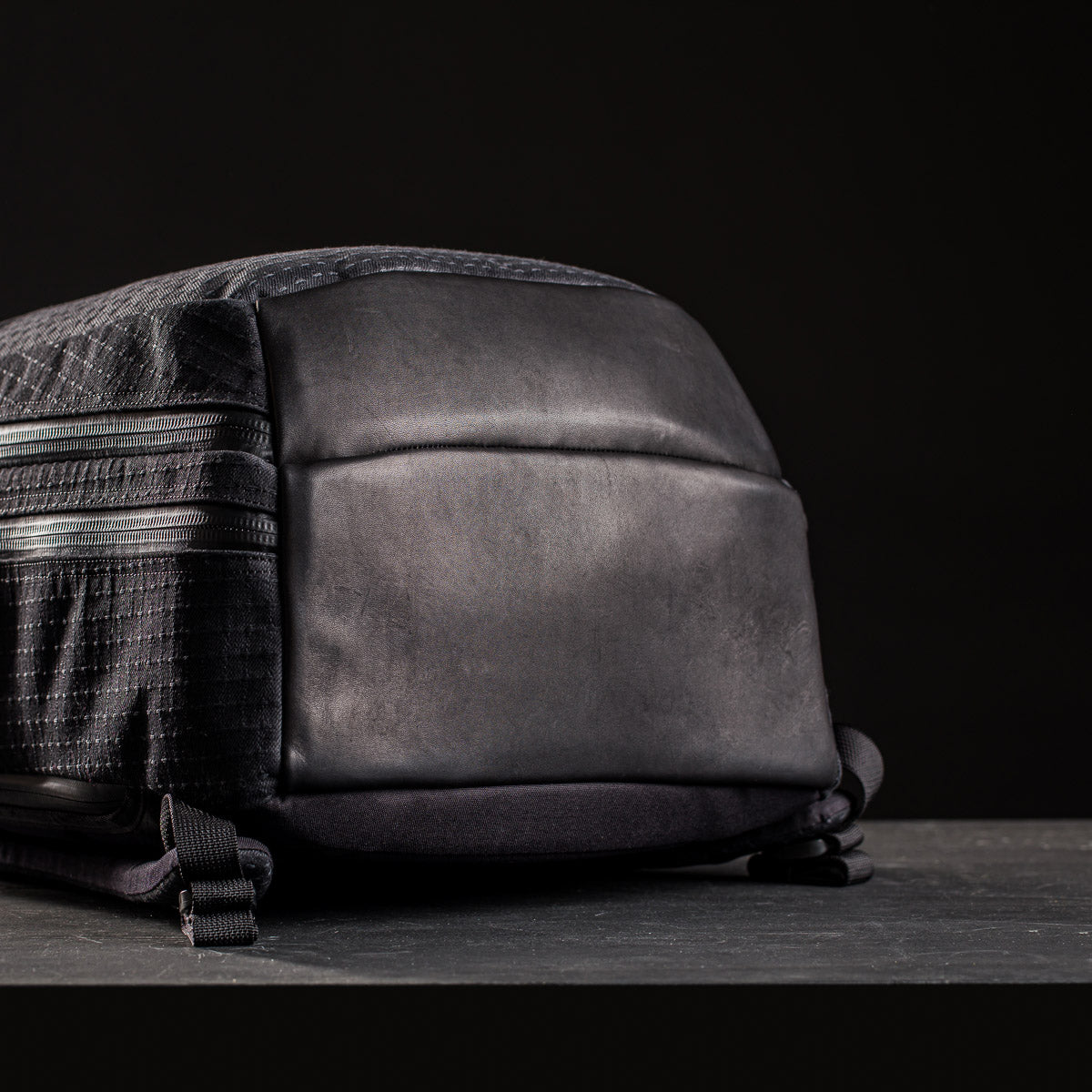 Close-up of the GR2 x Carryology - GRXC2 Samurai 2.0, a black backpack with a smooth leather bottom and zippered compartments, crafted from Japan Blue collaboration JPXDP denim, set against a dark background.