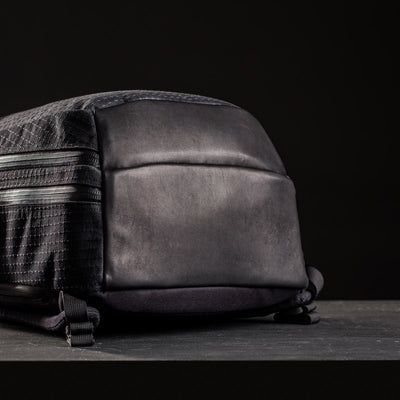 Close-up of the GR2 x Carryology - GRXC2 Samurai 2.0, a black backpack with a smooth leather bottom and zippered compartments, crafted from Japan Blue collaboration JPXDP denim, set against a dark background.