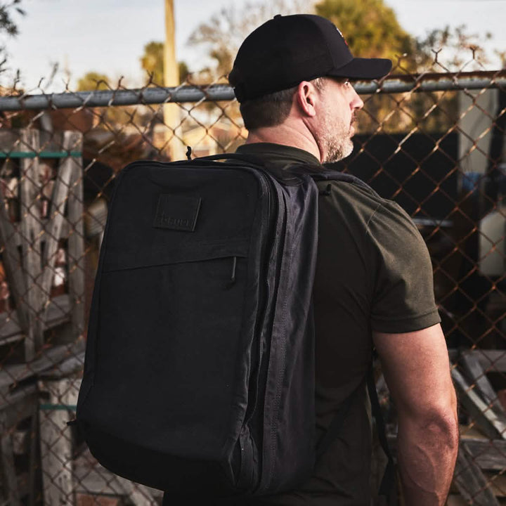 A person wearing a black cap and a large GR2 - Waxed Canvas by GORUCK stands by a chain-link fence. The setting appears to be outdoors with wooden pallets and scattered items in the background.