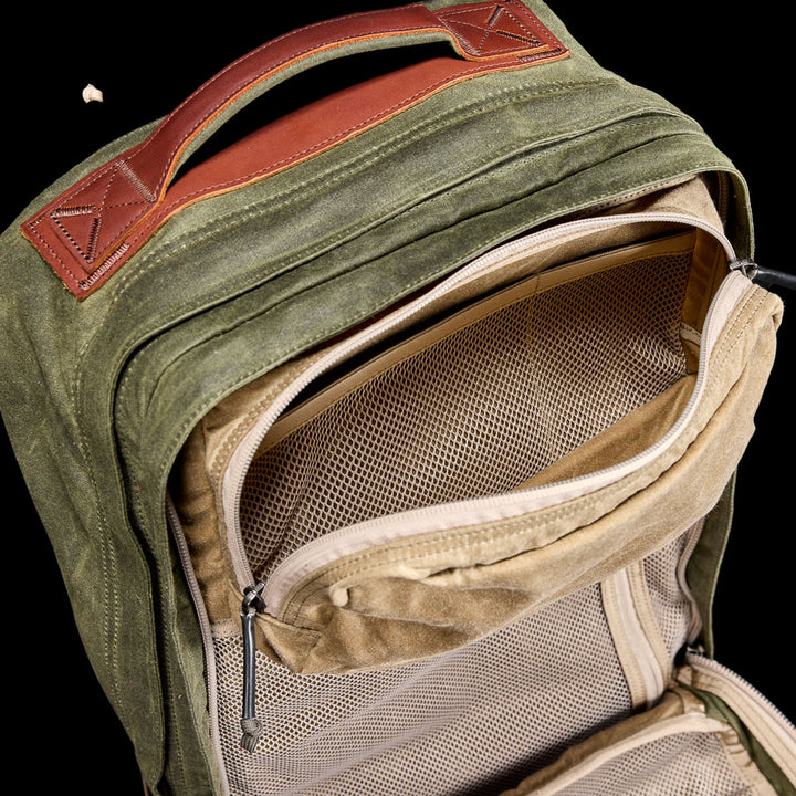 The GR2 - Waxed Canvas is a green backpack with a brown handle, shown partially unzipped to reveal mesh interior pockets against a black background—a durable travel ruck built for adventures.