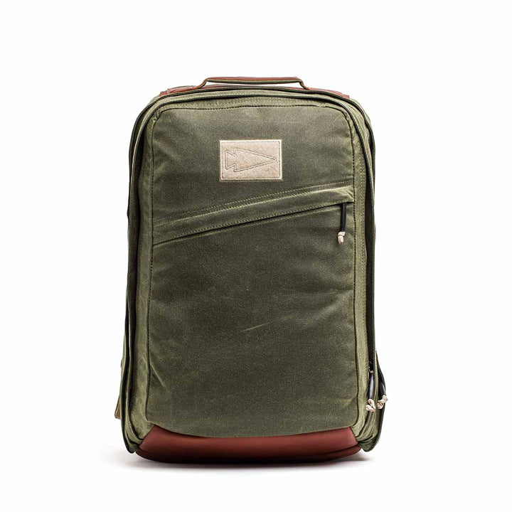 The GR2 - Waxed Canvas is an olive green backpack with a brown leather base, front zipper pocket, and beige patch. Made from durable duck canvas, it's perfect for daily use or travel and built for lasting style.