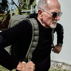 An older man in black attire and sunglasses walks outdoors by a white wall with the GR2 USA - Cordura travel backpack slung over his shoulder.