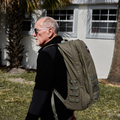 An older man wearing sunglasses and carrying the GR2 USA - Cordura khaki travel backpack walks outside near a white building and palm tree.