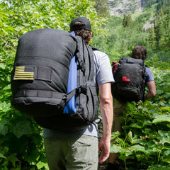 Two hikers rucking with large black GORUCK backpacks on a green forest trail in mountainous terrain