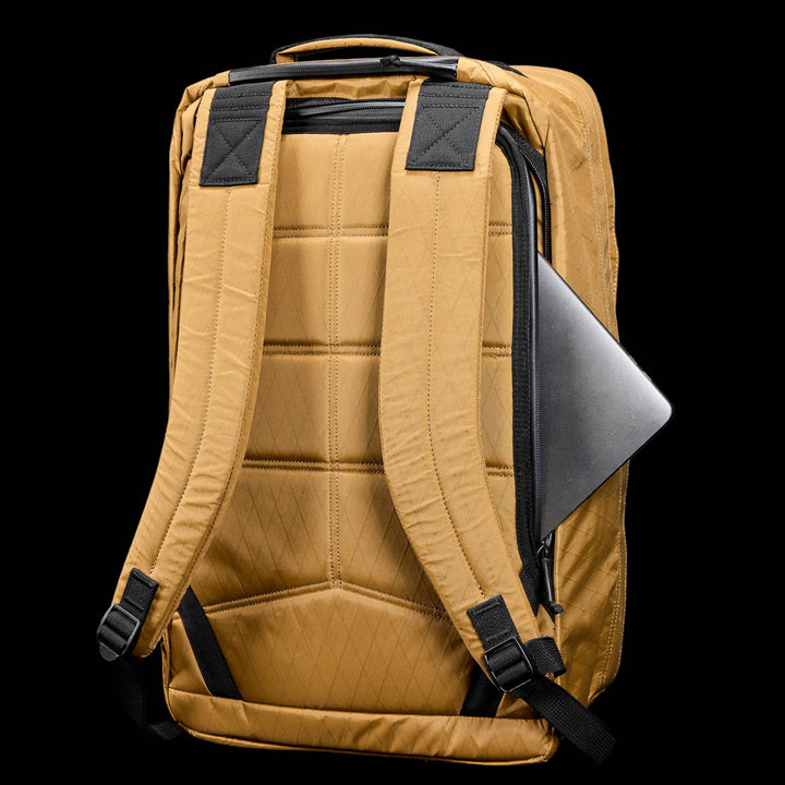 The GR2 X-PAC tan backpack with padded straps and AquaGuard zippers sits on a black background, featuring a laptop partially visible in its side zipper pocket.