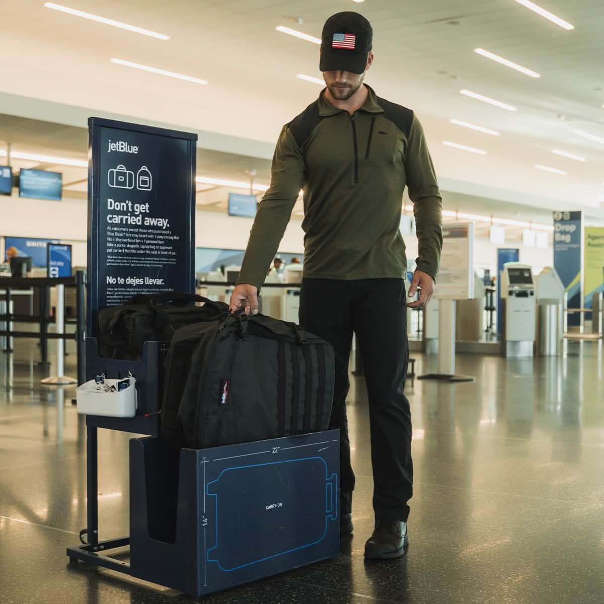 A person in a cap and casual attire is having difficulty fitting their GR3 - Dyneema carry-on backpack by GORUCK into a baggage sizer at an airport. A sign next to them reads, 