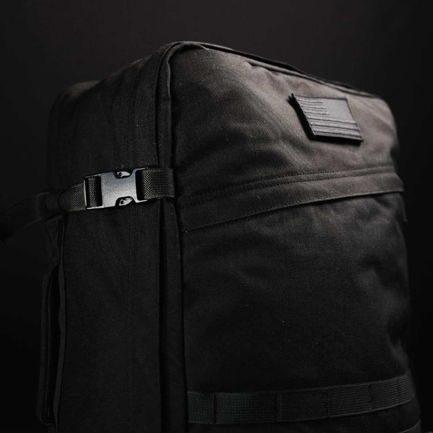 Close-up of a black backpack with buckles and a rectangular patch, set against a dark background.