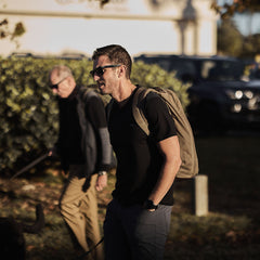 A man in sunglasses and a backpack sports the Men’s Commando Tee - Merino Wool, featuring reinforced shoulders, while walking outdoors with another person visible in the sunny background.