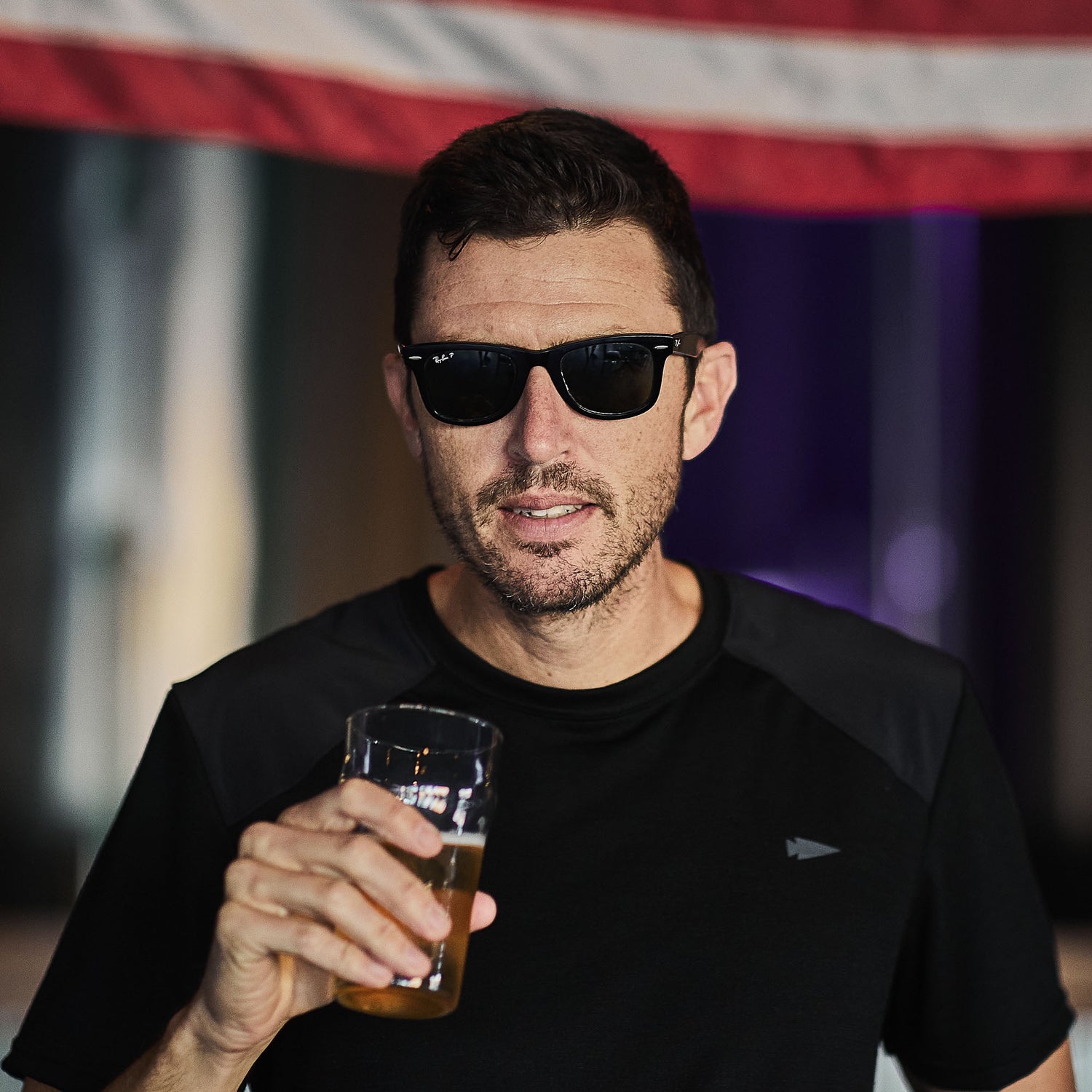 A man in sunglasses and the Men’s Commando Tee - Merino Wool with reinforced shoulders holds a beer, standing before an American flag.