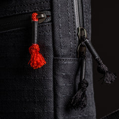Close-up of the GRenade x Carryology - GRXC2 Samurai 2.0, a black textured tactical sling with red and black tassel zipper pulls.