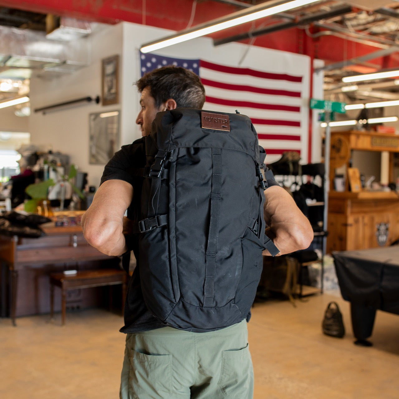 A man wearing a large black GR MONSTER Rucksack - PREORDER in a room with an American flag hanging on the wall.